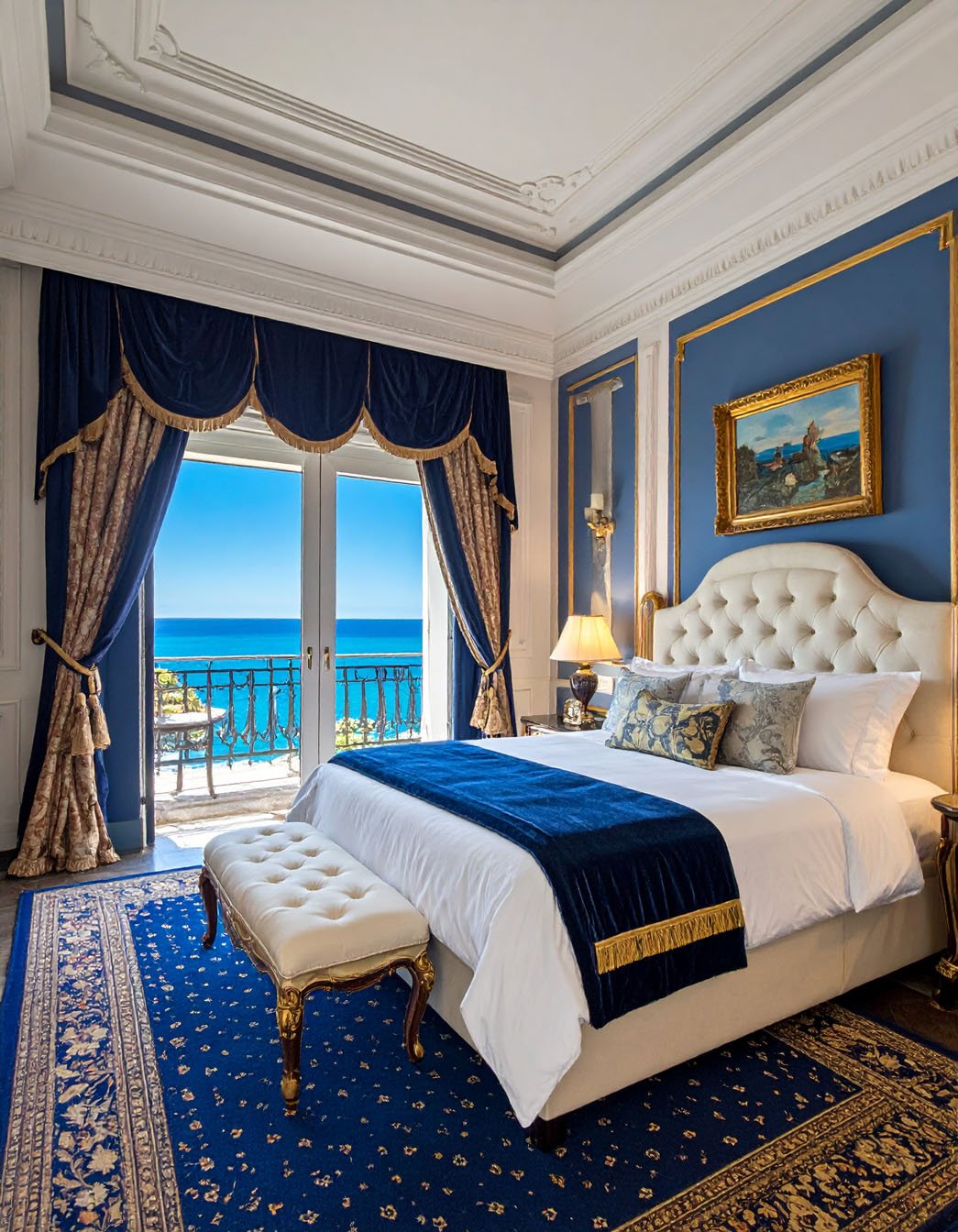 Chambre opulente avec murs bleus et dorures, lit blanc, vue mer, rideaux somptueux