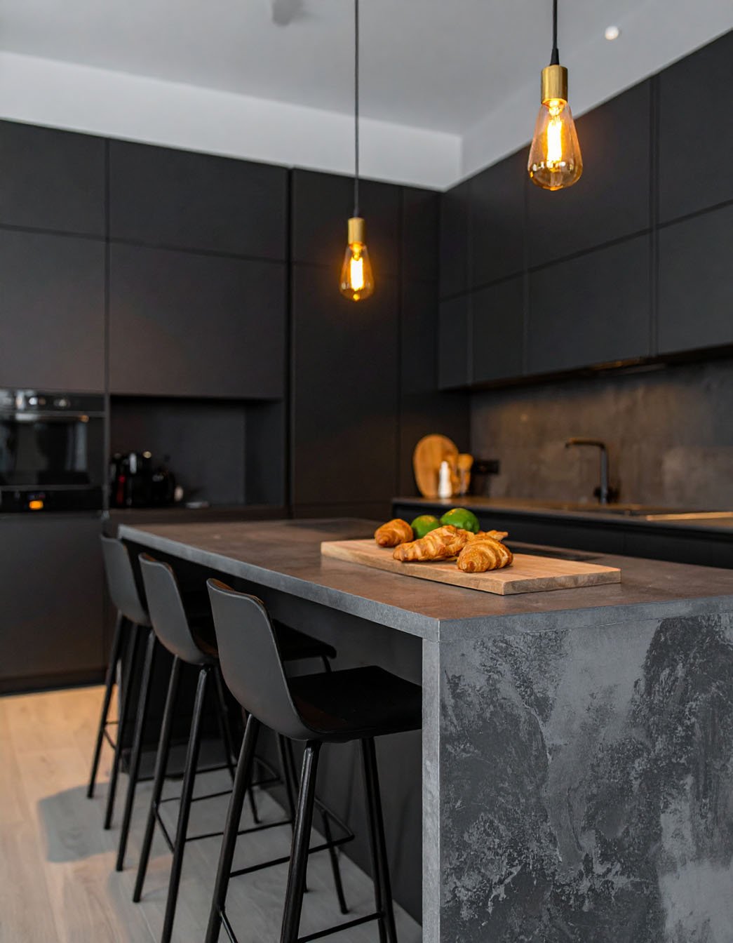 Cuisine noire avec îlot effet béton ciré, tabourets noirs et suspensions à ampoules Edison