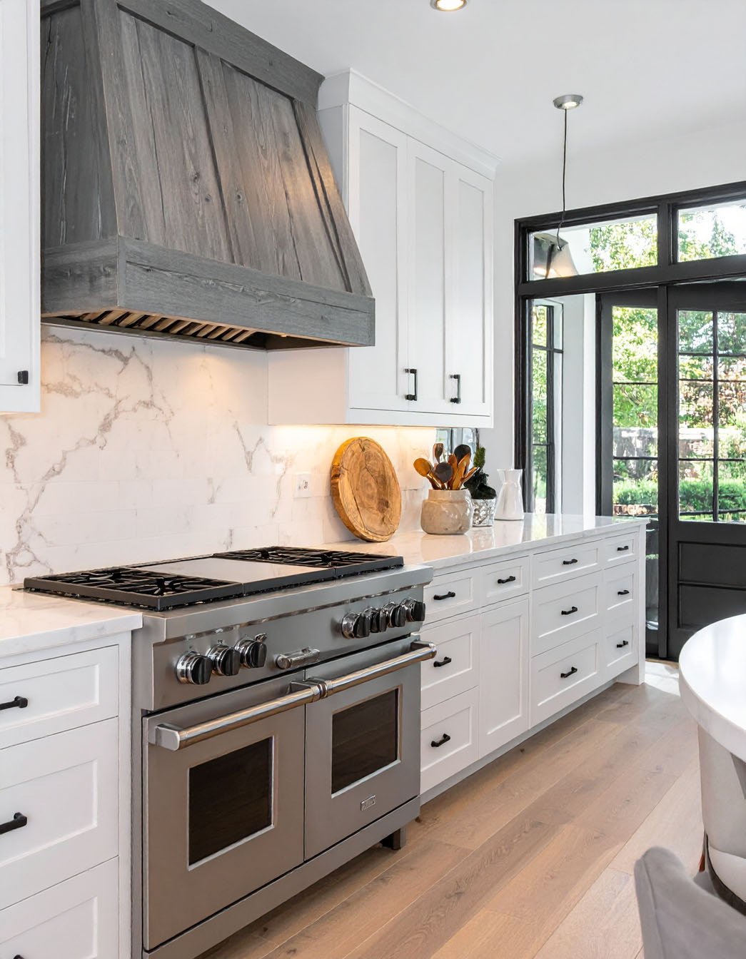 Cuisine blanche avec hotte en bois grisé, piano de cuisson inox et porte noire vitrée