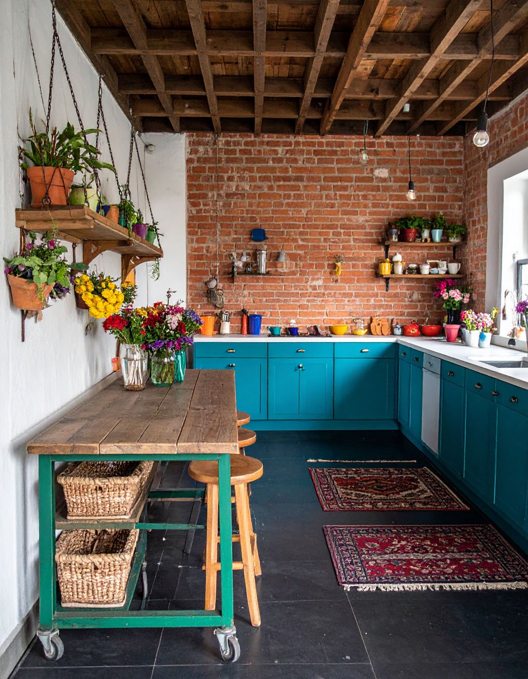 Cuisine bohème avec meubles bleu canard, mur en briques, table en bois et plantes suspendues