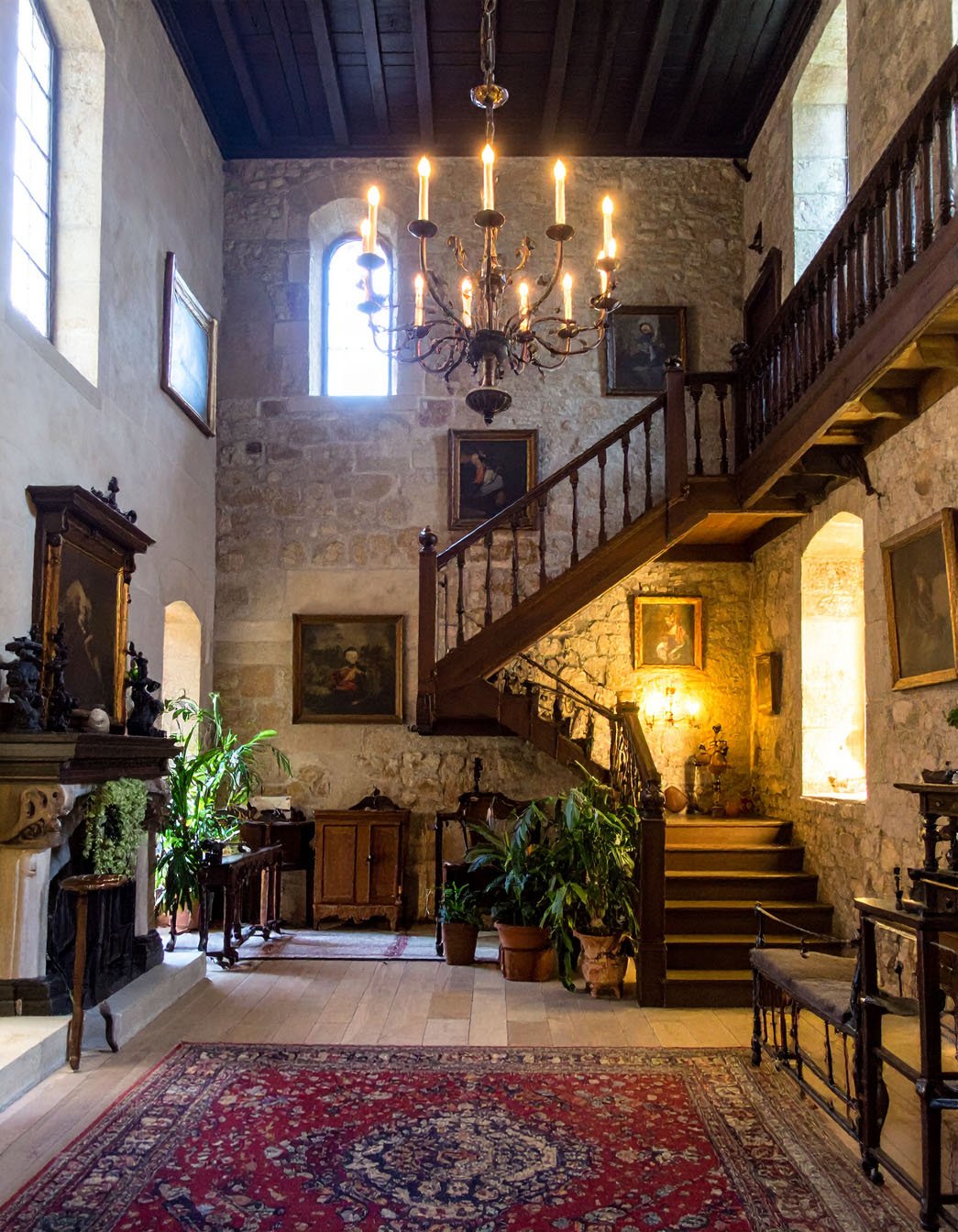 Hall d'entrée de château avec murs en pierre, escalier en bois et lustre à bougies