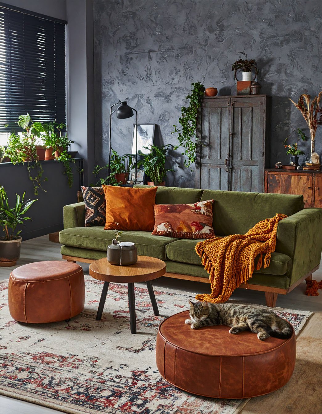 Salon avec mur texturé gris foncé, canapé en velours vert olive, poufs en cuir et chat tigré