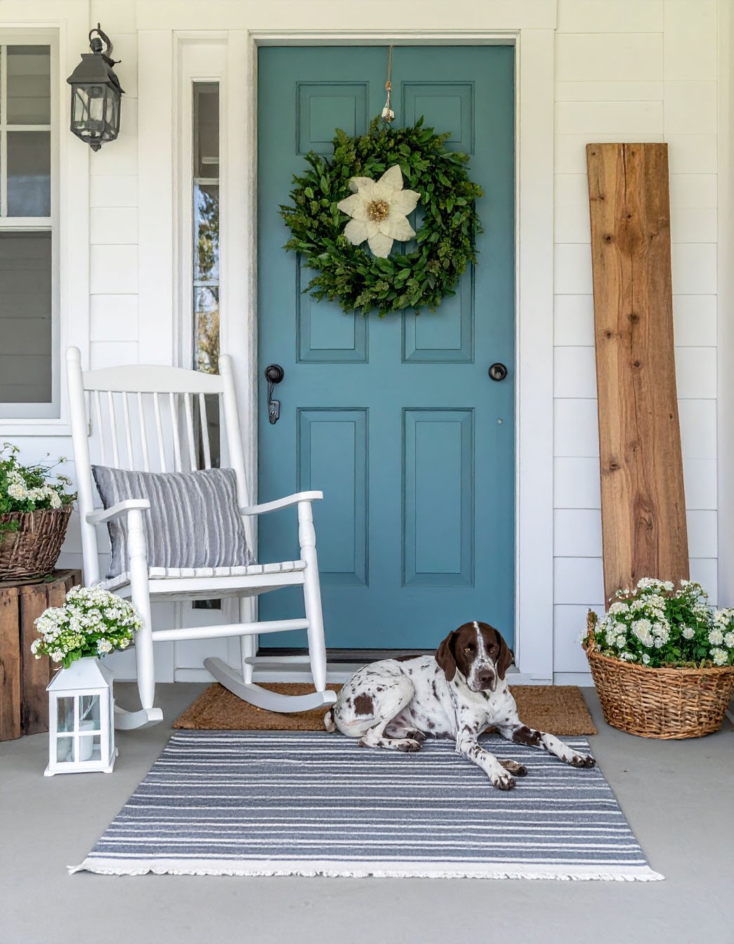 Porte d'entrée bleu-vert avec couronne, fauteuil à bascule blanc, chien tacheté couché