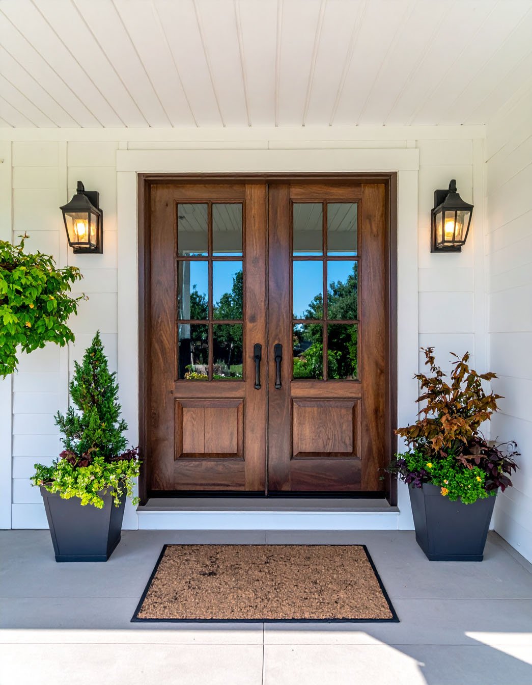 Double porte d'entrée en bois vitrée, encadrée d'appliques et de grands pots de fleurs
