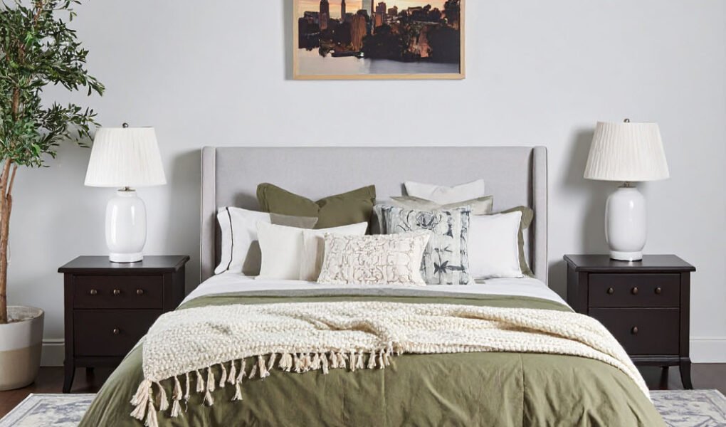 Chambre avec lit gris, literie vert olive et plaid crème, tapis à motifs et tableau de ville
