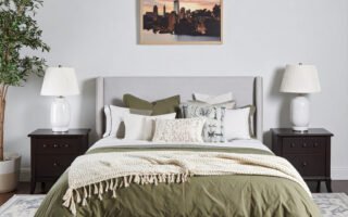 Chambre avec lit gris, literie vert olive et plaid crème, tapis à motifs et tableau de ville
