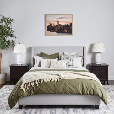 Chambre avec lit gris, literie vert olive et plaid crème, tapis à motifs et tableau de ville