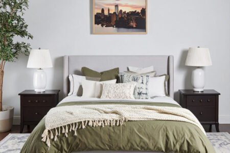 Chambre avec lit gris, literie vert olive et plaid crème, tapis à motifs et tableau de ville