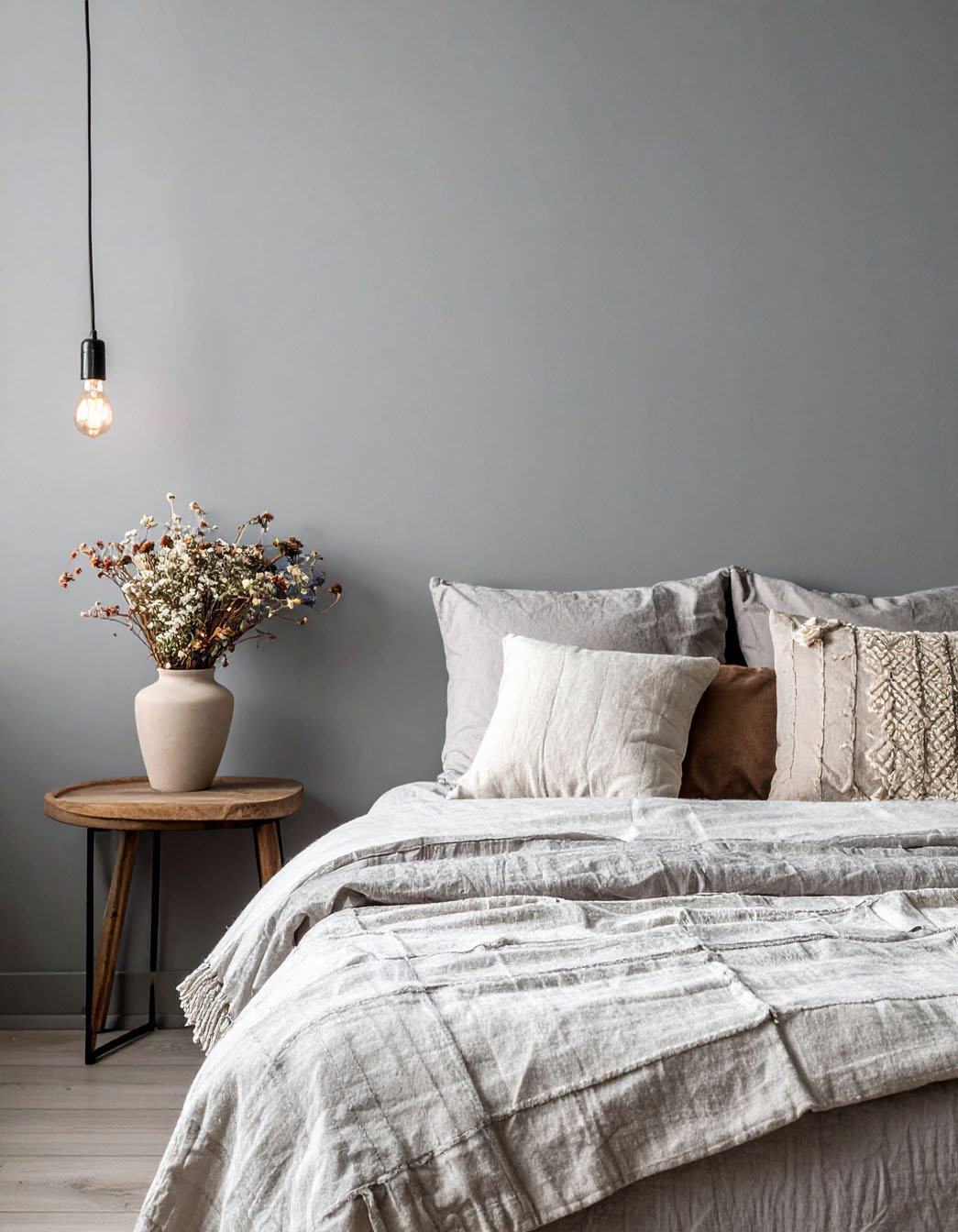 Chambre aux murs gris avec lit, coussins texturés et table d'appoint en bois avec fleurs séchées