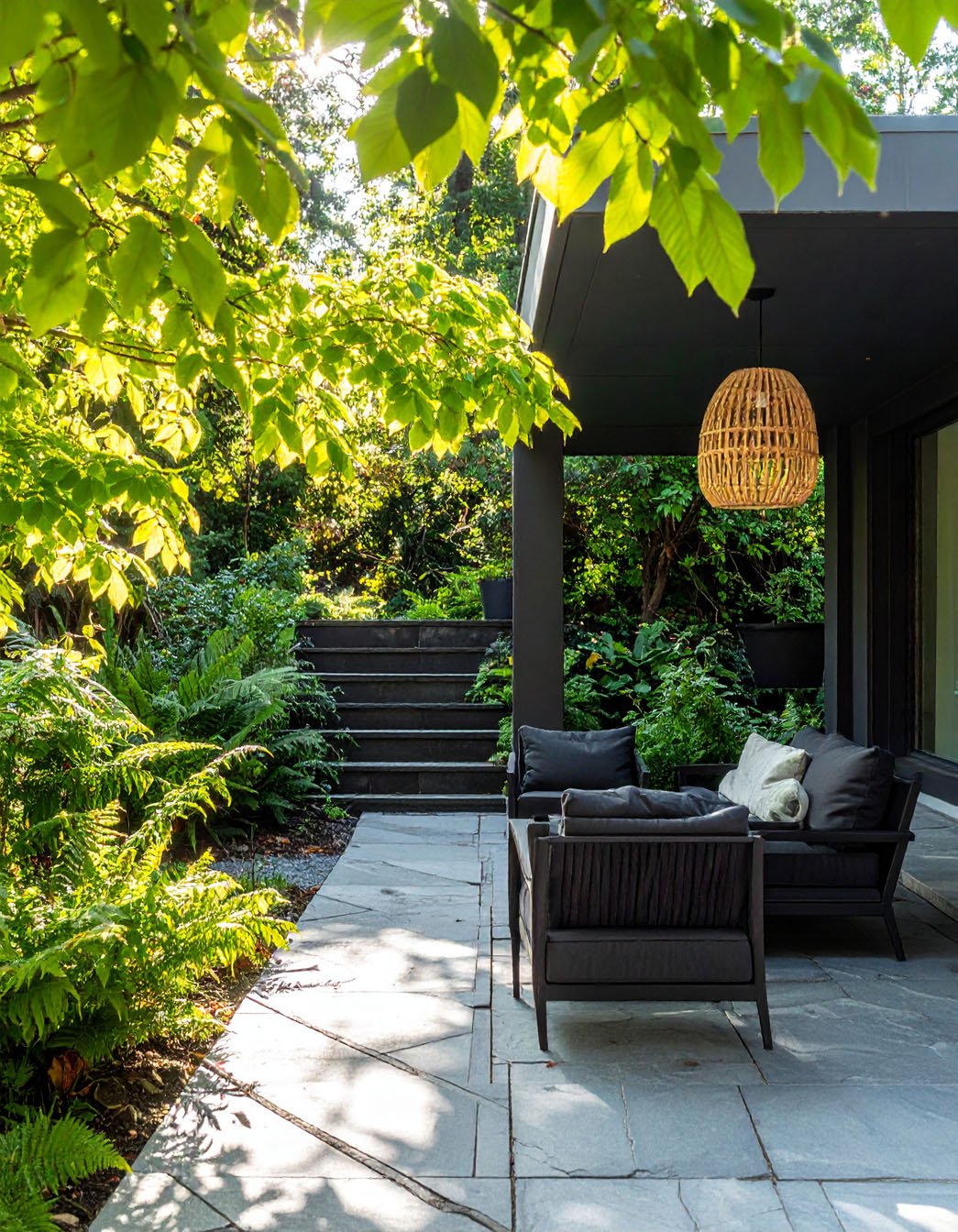 Terrasse ombragée avec salon de jardin noir, suspension en rotin et végétation luxuriante