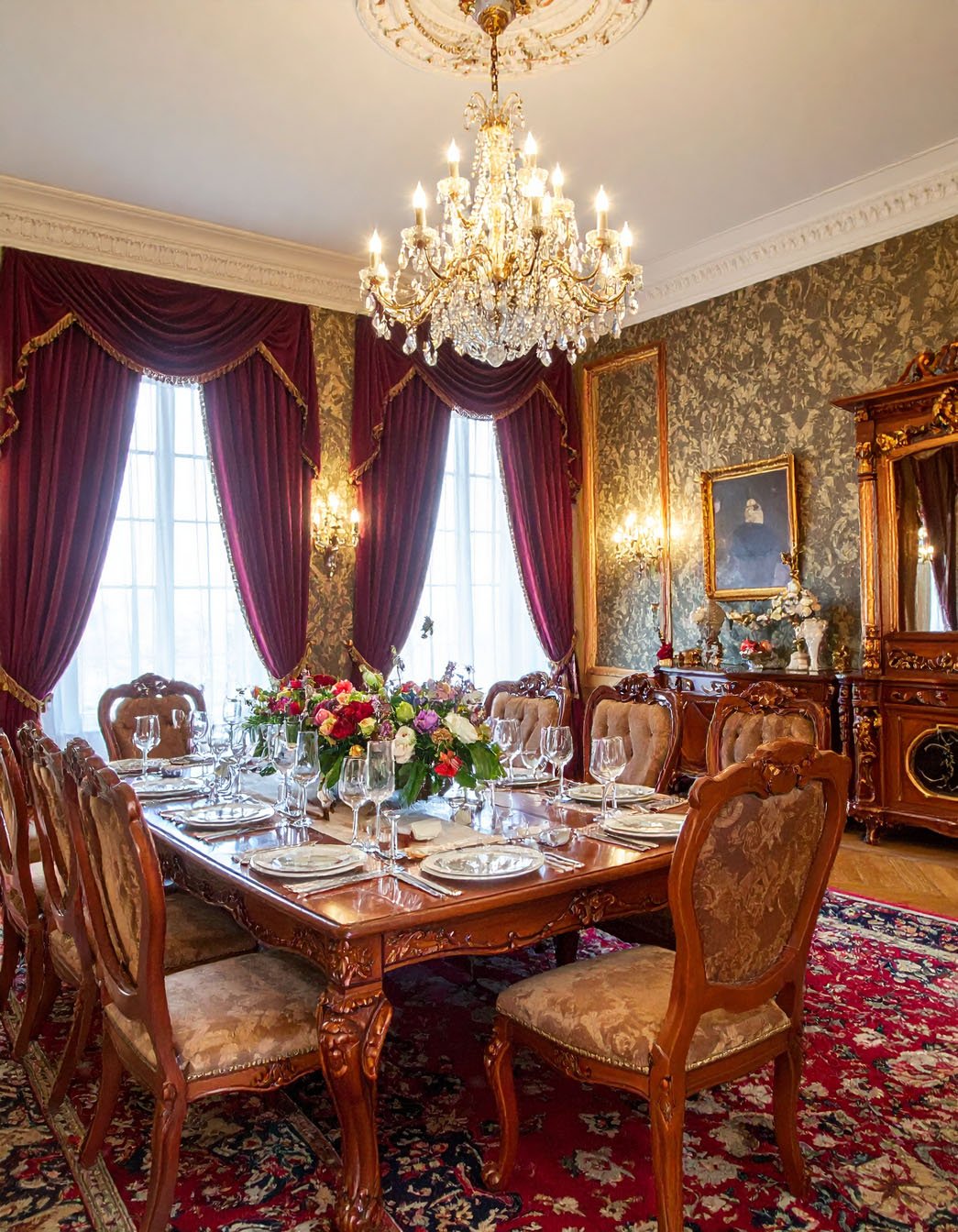 Salle à manger opulente avec table dressée, chaises style ancien, lustre en cristal et rideaux carmin