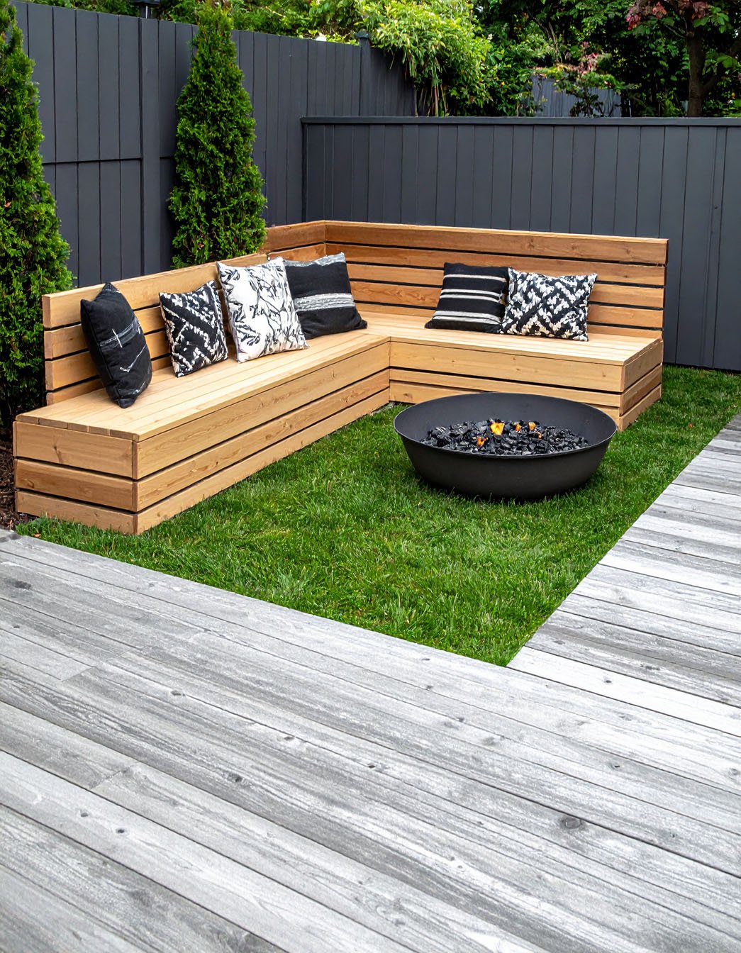 Terrasse en dalles avec banquette d'angle en bois et coussins blancs, entourée de verdure
