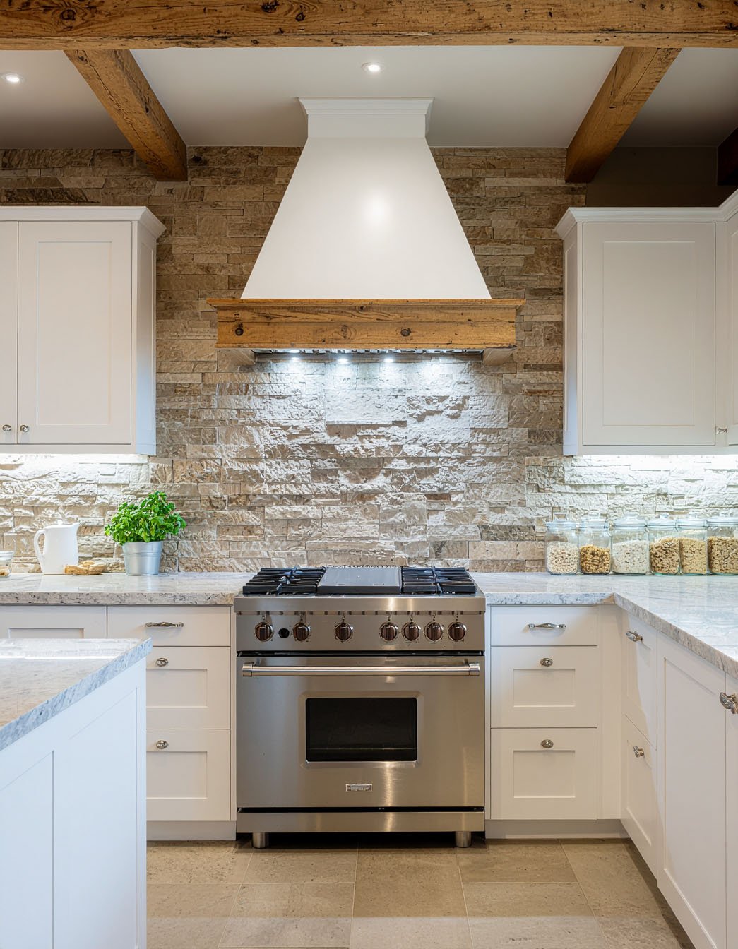Cuisine avec meubles blancs, mur et hotte en pierres, poutres en bois apparentes